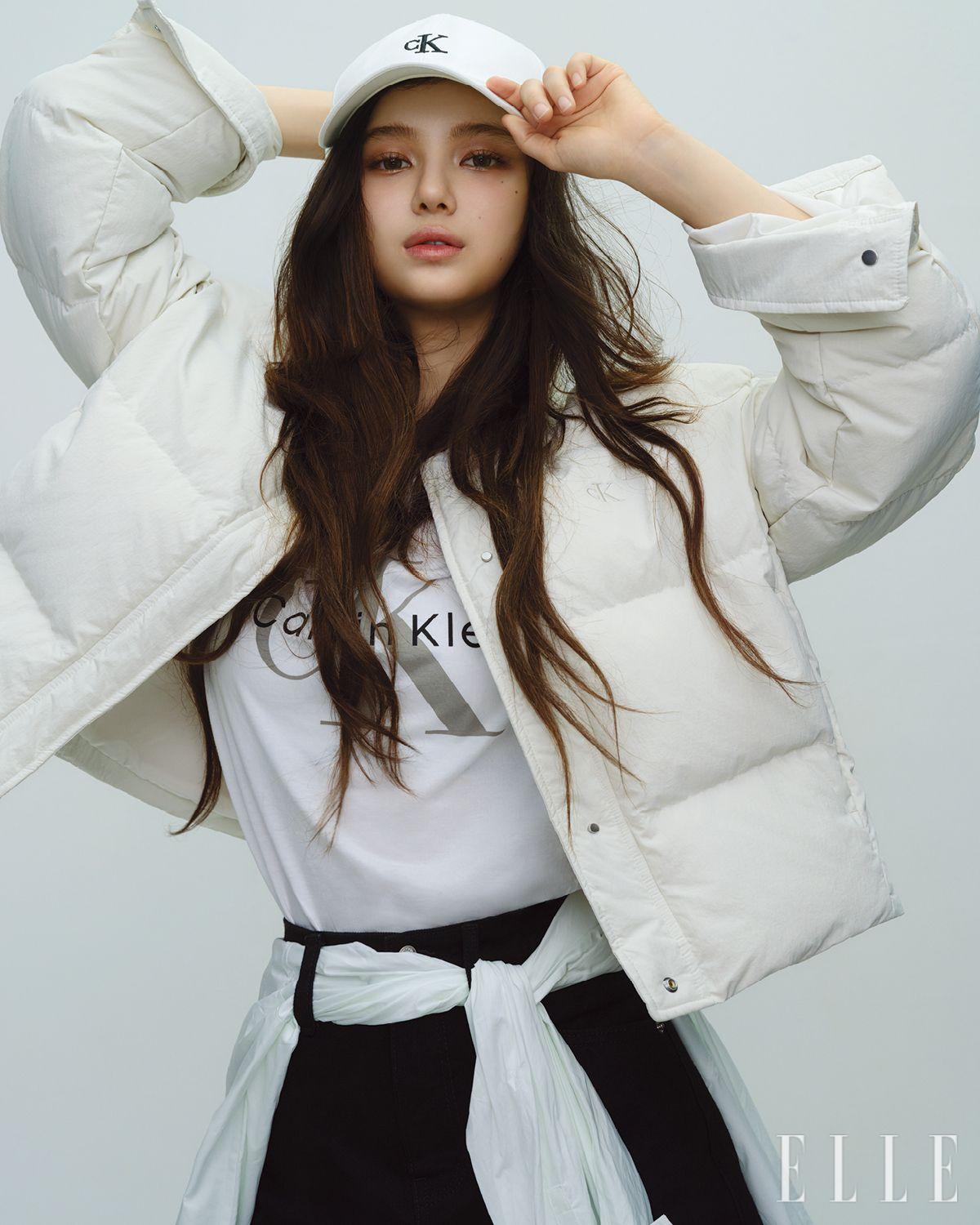 다니엘이 입은 화이트 퀼티드 다운 재킷과 크루넥 디자인의 로고 티셔츠, 데님 하이라이즈 미니스커트, 허리에 두른 코튼 포플린 셔츠, 로고 볼 캡은 모두 Calvin Klein Jeans. 