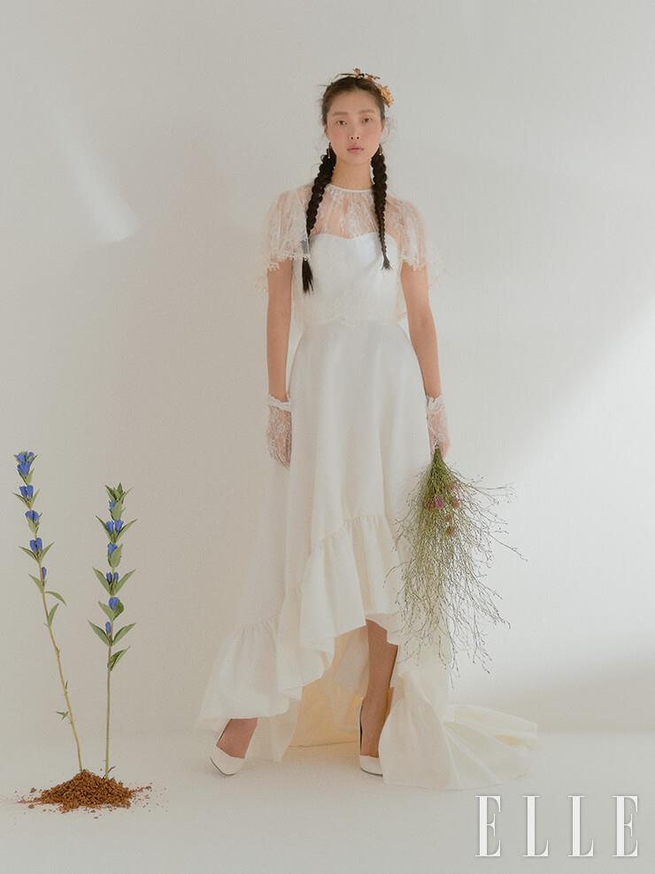 케이프 스타일의 레이스 톱과 페플럼 디테일의 언밸런스 드레스는 모두 Halfpenny London by Soyoo Bridal. 헤어 오브제와 레이스 글러브는 모두 The Queen Lounge. 이어링은 Ros&eacute;eD'or. 화이트 펌프스는 Stuart Weitzman.