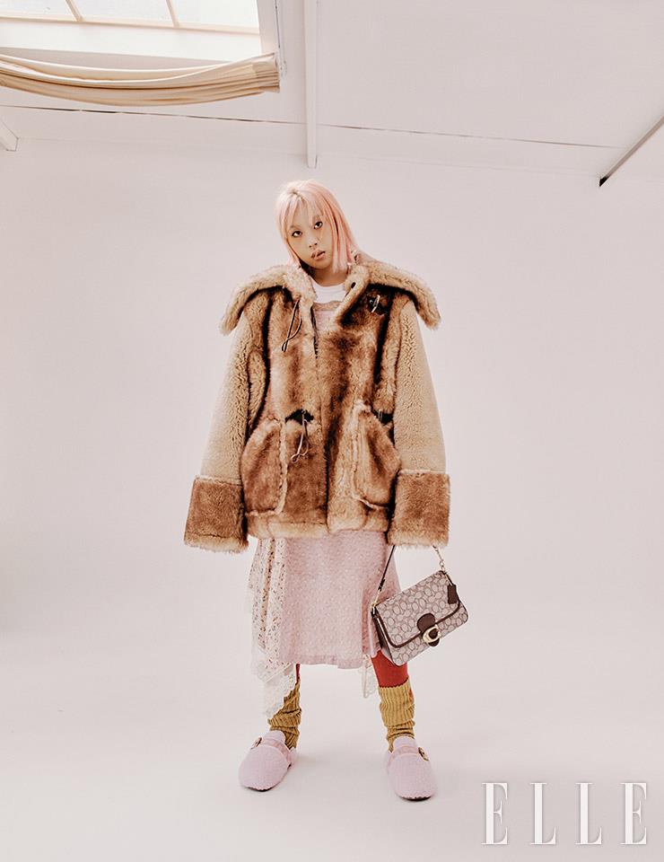 오버사이즈 시어링 코트는 Coach x Schott NYC. 퀼팅 장식의 프린트 티셔츠와 란제리 스커트 위에 레이어드한 에이프런 스커트, 시그너처 자카르의 소프트 태비 숄더백, 핑크 컬러 시어링 크로그, 레깅스, 양말은 모두 Coach.