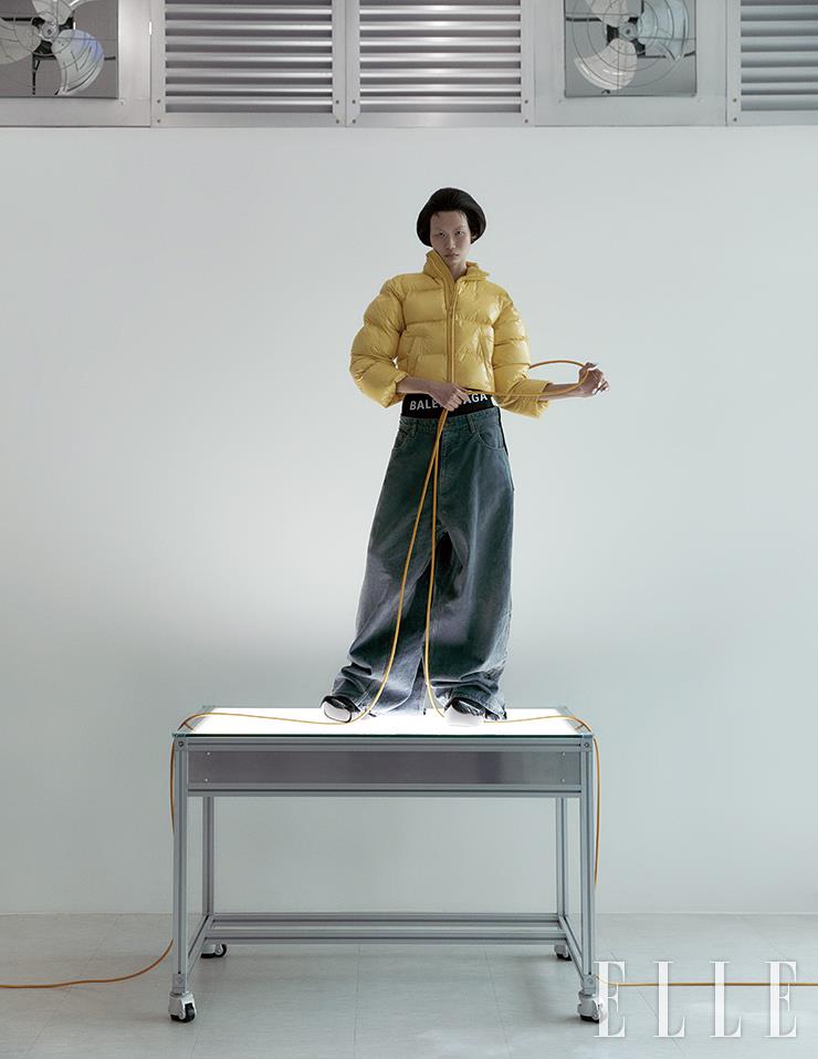 옐로 컬러 푸퍼 패딩과 빅 로고 브리프, 에이프런 스타일의 데님 팬츠, 글로시한 크로그는 모두 Balenciaga.